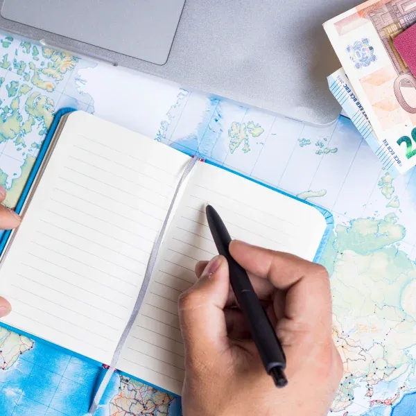 A man writes in a notebook on a map, with a pen and laptop, focused on travel financing.