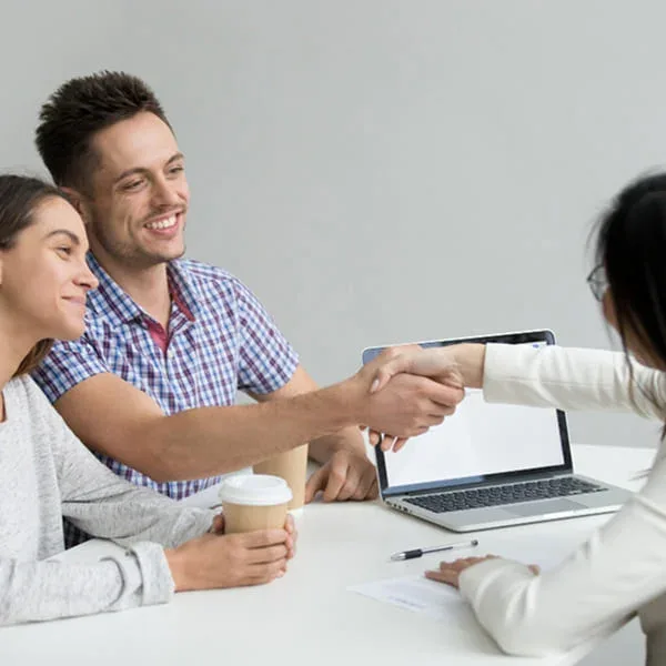 A man and woman shaking hands at a table with a laptop open between them, indicating a business agreement.