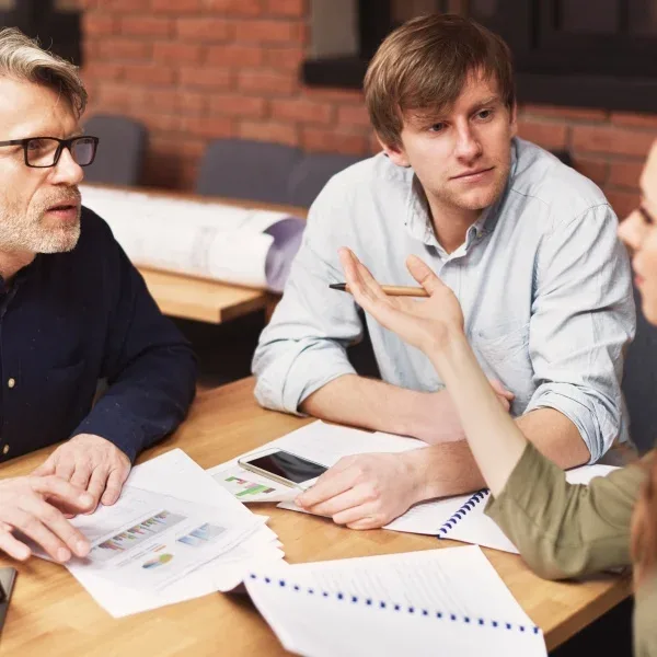 Trois personnes assises à une table, examinant des documents et utilisant un ordinateur portable lors d'une discussion collaborative.