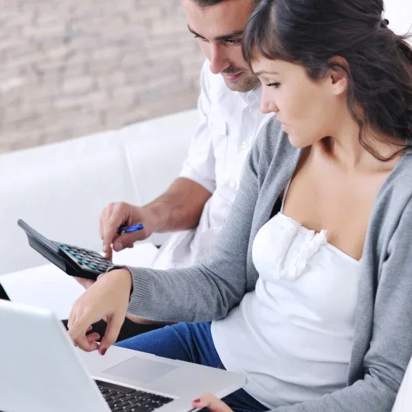 A man and woman sit together on a couch, focused on a laptop placed on their laps.