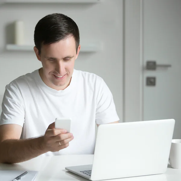 A man sitting at a table, using a laptop and a cell phone, focused on his work.