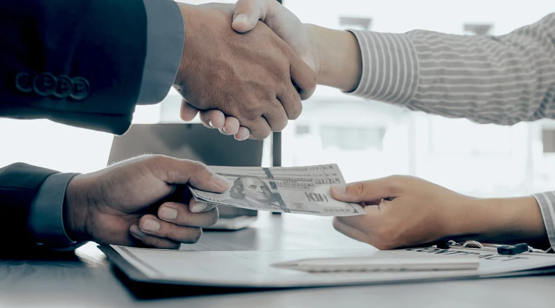Two people shaking hands over a stack of cash on a desk, symbolizing a business agreement or transaction.