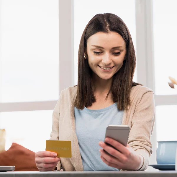A woman holds a credit card in one hand and a smartphone in the other, appearing engaged in a transaction.